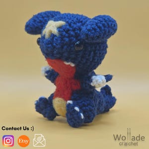 May include: A crocheted plush toy depicting a cartoon character. The toy is predominantly dark blue, featuring a red chest, white teeth, and a yellow star on its head. It has black eyes and white detailing on its arms and feet.