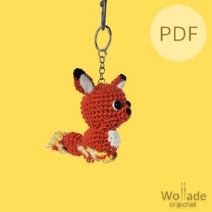 Op de afbeelding: Een gehaakte vos sleutelhanger met een rood-gele staart, hangend aan een zilveren sleutelring. De vos heeft zwarte ogen en een witte buik. De achtergrond is geel. De tekst "PDF" is zichtbaar in de rechterbovenhoek van de afbeelding.