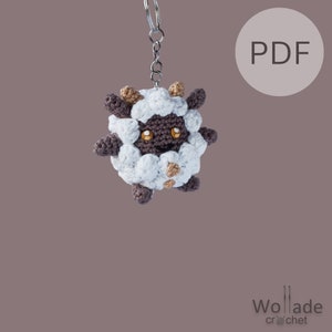 Op de afbeelding: Een wit en bruin gehaakt amigurumi-speelgoed van een schaap met een sleutelhanger. Het speelgoed heeft een bruin gezicht met twee oranje ogen en een kleine glimlach. Het speelgoed hangt aan een zilveren sleutelhanger. De achtergrond is lichtbruin. De tekst "PDF" staat in een witte cirkel aan de rechterkant van de afbeelding.