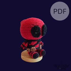 Op de afbeelding: Een gehaakte Deadpool amigurumi-pop die op een houten voetstuk zit. De pop is rood en zwart met een zwart oogmasker. De pop draagt een rood en zwart pak met een zwarte riem. De pop zit op een houten voetstuk. De afbeelding bevat de tekst "PDF".
