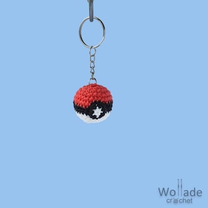 Op de afbeelding: Een handgemaakte gehaakte sleutelhanger met een rood, zwart en wit balontwerp. De bal heeft een stervormig detail en is bevestigd aan een zilveren sleutelring. Een uniek accessoire.