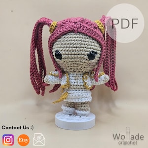 Op de afbeelding: Een handgemaakte gehaakte pop met een roze en gouden hoed en lange roze vlechten. De pop draagt een witte outfit met gouden accenten en staat op een witte basis. De pop heeft grote zwarte ogen. De tekst "PDF" is inbegrepen.