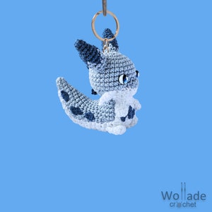 Puede incluir: Un llavero de amigurumi de crochet azul y blanco con forma de criatura mítica con cola larga y ojos grandes. El llavero está unido a un anillo de llave de metal dorado.