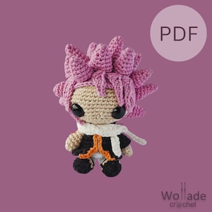 Op de afbeelding: Een roze en beige gehaakte amigurumi pop met zwarte ogen en een witte sjaal. De pop draagt een zwart en oranje outfit. De afbeelding heeft het woord "PDF" in een witte cirkel op een roze achtergrond.