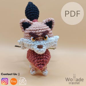 May include: A crochet fox figurine featuring a tan head with black-tipped ears, a white muzzle, and a dark brown nose. The fox has orange eyes, dark brown eyebrows, and a black tuft on top. It wears a reddish-brown chest piece and has white paws.