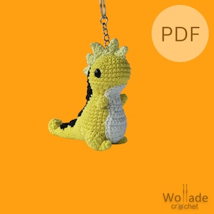 May include: A yellow and black crocheted dinosaur keyring with a white belly and black eyes. The keyring is attached to a silver chain. The image also includes the word "PDF" in a circle.