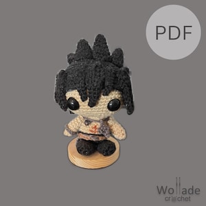May include: A crocheted doll with black hair and large black eyes, standing on a small wooden base. The doll has a tan body with dark accents and is wearing a brown belt. The image includes a "PDF" graphic and the "Wollade crochet" logo.