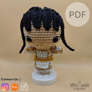 Op de afbeelding: Een handgemaakte gehaakte pop met zwart haar en grote zwarte ogen. De pop draagt een crèmekleurige outfit met gouden accenten. De pop staat op een witte basis. De afbeelding bevat de tekst "PDF" en "Wollade crochet".