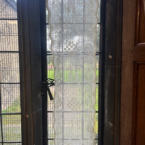 Puede incluir: Una cortina de encaje blanco cuelga en una ventana con vista a un césped verde y un edificio de piedra.
