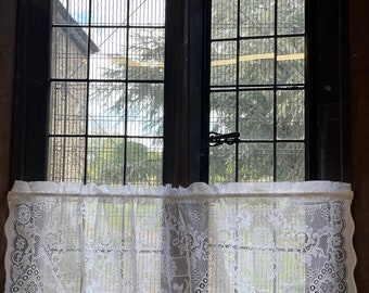May include: White lace valance with a scalloped edge hangs in front of a window with a black metal grid. The window is divided into multiple panes and looks out onto a green tree.