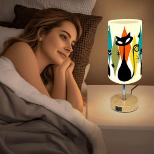 May include: A retro-style table lamp with a cylindrical shade featuring a black cat silhouette and colorful diamond shapes. The lamp has a wooden base and a chrome stem. The design includes orange, teal, and white accents, creating a mid-century modern aesthetic.