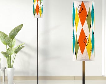 May include: A floor lamp with a tall, thin black metal pole and a cylindrical fabric shade. The shade features a repeating pattern of orange, yellow, teal, and olive green diamonds interspersed with small black starburst and diamond line icons.