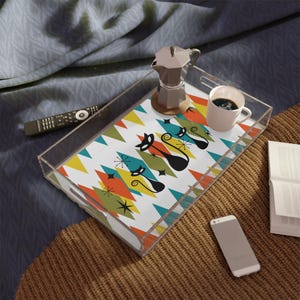 May include: A clear acrylic serving tray with a colorful mid-century modern design. The tray features a pattern of diamonds and triangles in orange, yellow, teal, and green, with black cat silhouettes. A coffee pot, a cup of coffee, and a remote control are on the tray.