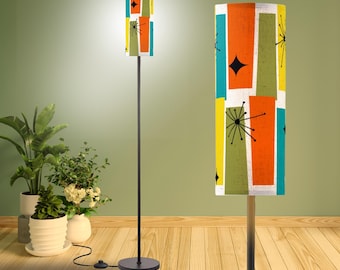 May include: A floor lamp with a tall, cylindrical shade featuring a colorful pattern of orange, teal, yellow, and olive green rectangles accented by black starburst and diamond shapes. The lamp has a thin, black metal pole and a circular base.