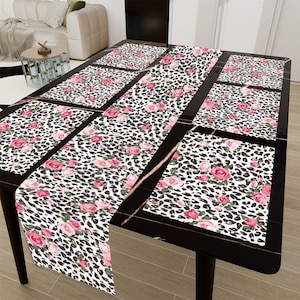 May include: A set of six black and white leopard print placemats with pink roses, and a matching black and white leopard print table runner with pink roses. The placemats and table runner are on a black table with a gold trim.