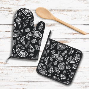 May include: Black and white paisley oven mitt and pot holder set. The kitchen textiles feature a classic bandana pattern. A wooden spoon rests nearby on a white-washed wooden surface.