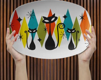 May include: An oval serving platter with a retro design. The white platter features black cat silhouettes and starbursts against a backdrop of colourful diamonds in red, yellow, teal, orange, and green. A fun, mid-century modern design.