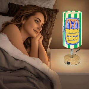 May include: A bedside lamp with a cylindrical lampshade featuring a sardine can design. The can is yellow with blue fish and the text "WILD CAUGHT Sardines". The lamp has a wooden base and a chrome stem.
