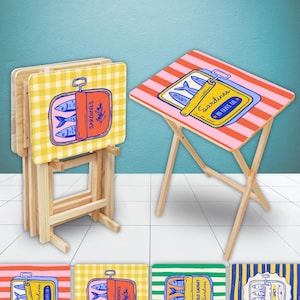 May include: A set of wooden folding snack tables with light wood legs. One table is open, featuring a red and pink striped top with a sardine tin graphic labeled Sardines In Olive Oil. Others are folded, showing yellow gingham and sardine prints.