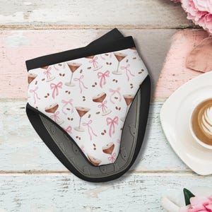 May include: A black and gray oven mitt with a white surface featuring a pattern of espresso martinis, pink bows, and coffee beans. The mitt is on a light blue and pink wooden surface, with a cup of coffee and flowers in the background.