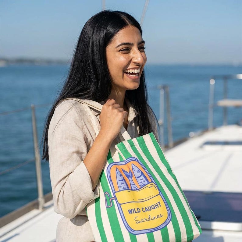 May include: A green and white striped tote bag featuring a sardine can illustration with the text "WILD CAUGHT Sardines." The bag is held by a person on a boat, with the ocean in the background.