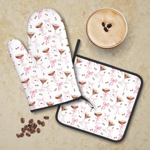 May include: A set of two kitchen accessories featuring a white background with a repeating pattern of pink bows, coffee beans, and brown martini glasses. The set includes a white oven mitt and a white pot holder.
