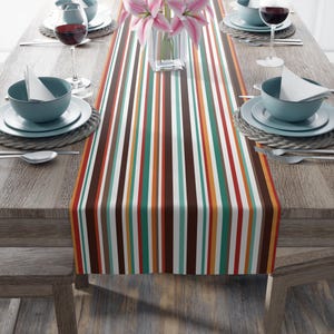 May include: A dining table set for a meal, featuring a colorful striped table runner. The runner has vertical stripes in brown, red, orange, teal, and white. The table is set with teal bowls, white plates, wine glasses, and silverware.