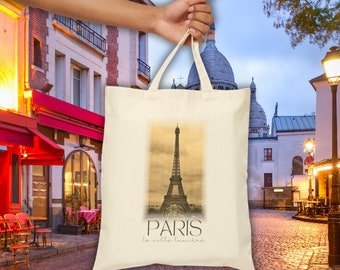 Könnte beinhalten: Eine beige Tragetasche mit einem Vintage-Bild des Eiffelturms und dem Text "PARIS la ville lumière" auf der Vorderseite. Die Tasche wird von einer Hand vor dem Hintergrund einer Pariser Straßenszene gehalten.
