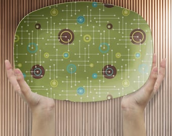 May include: Oval-shaped serving platter with a retro design. The platter features a light green background with a pattern of brown, blue, and yellow circles and white lines. The platter is held by two hands.