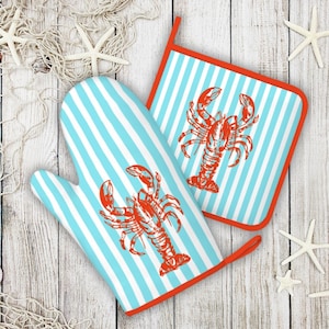May include: A blue and white striped oven mitt and pot holder set. Each item features a red lobster design. The edges of the pot holder and the trim of the oven mitt are red. The set is on a white wooden surface.