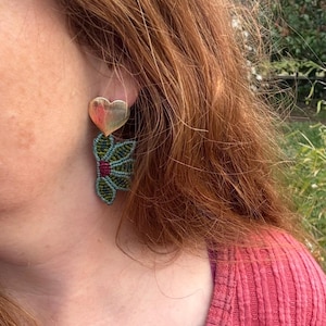May include: Earrings featuring a gold heart-shaped top and a green and teal macrame flower design. These earrings are a unique accessory, ideal for adding a touch of bohemian flair to any outfit. The earrings are a handmade item.