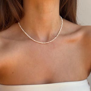 May include: Delicate necklace composed of small, round, white pearls. The necklace is worn around the neck, resting on the collarbone. The pearls are strung together closely, creating a continuous strand of white. The background is a neutral tone.