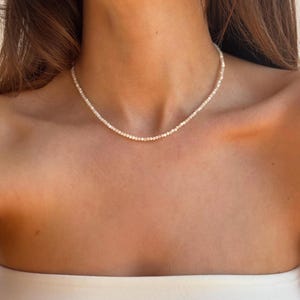 May include: Delicate necklace featuring small, round, off-white beads. The necklace is strung closely together, creating a subtle, elegant look. The beads appear to be uniform in size and shape, forming a continuous strand. This necklace is a classic piece of jewelry.