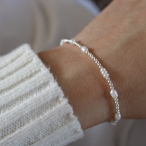 May include: A delicate bracelet featuring small white pearls and silver beads. The bracelet is worn on a person's wrist.