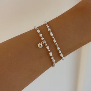 Könnte beinhalten: Zwei Armbänder mit Perlen. Ein Armband hat weiße und silberne Perlen mit einem kleinen Muschel-Anhänger. Das andere Armband besteht aus weißen und silbernen Perlen. Die Armbänder werden am Arm einer Person getragen.