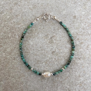 May include: A delicate bracelet made with small green gemstone beads and a single white pearl accent. The bracelet has a silver clasp.