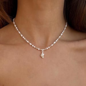 Pode incluir: Um colar delicado com pequenas contas brancas intercaladas com contas prateadas. Um pequeno pingente de concha prateada pende do centro. O colar foi projetado para ser usado ao redor do pescoço.