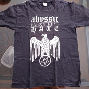 May include: Dark gray t-shirt with white graphic text and a stylized eagle design. The text reads "abyssic", "HATE", and other symbols. The eagle is centered over a pentagram.