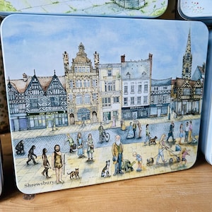 May include: Rectangular tin featuring a watercolour illustration of Shrewsbury, England. The image depicts a street scene with buildings, people, and the text "Shrewsbury". Other tins with similar illustrations are visible.