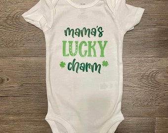May include: White baby onesie with green text that reads "mama's lucky charm" with shamrock symbols on either side of the word "charm".