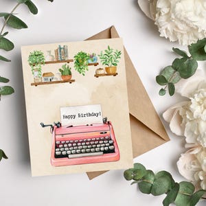 May include: A birthday card featuring a pink typewriter with the text "Happy Birthday!". The card has a watercolor illustration of shelves with potted plants and books. A brown envelope is behind the card.