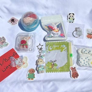 May include: A collection of small toys and accessories, including a fairy figurine, a keychain with a dog charm, and various stickers. The items are arranged on a white surface. A red card with the text "Fairy / Butterfly Wings" is also present.