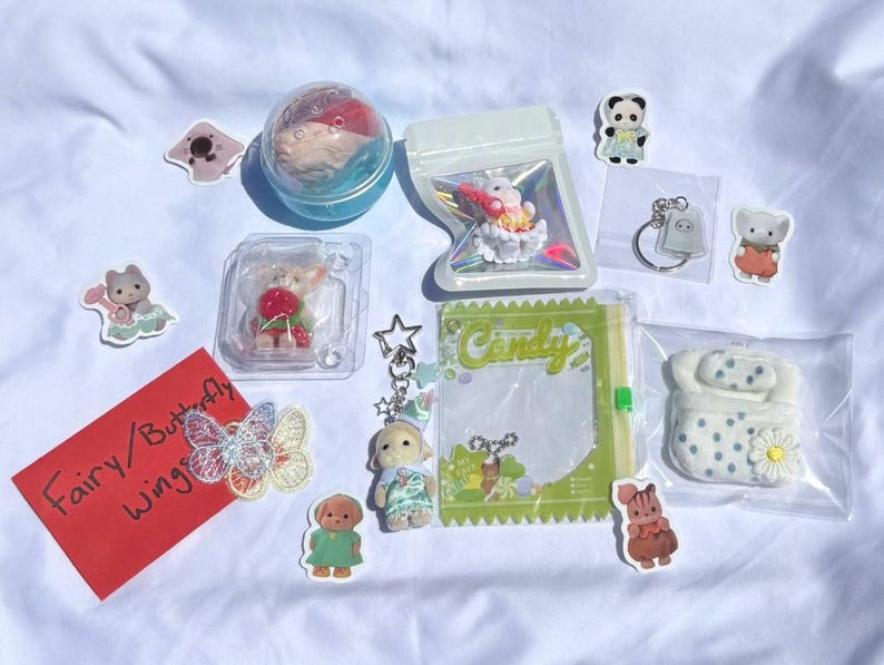 May include: A collection of small toys and accessories, including a fairy figurine, a keychain with a dog charm, and various stickers. The items are arranged on a white surface. A red card with the text "Fairy / Butterfly Wings" is also present.