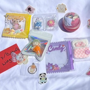 May include: A collection of miniature toys and stickers, including a bear in a yellow candy bag, a doll, a bed, and a butterfly. The items are arranged on a white surface.