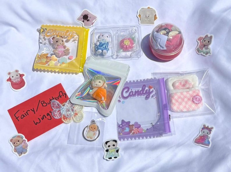 May include: A collection of miniature toys and stickers, including a bear in a yellow candy bag, a doll, a bed, and a butterfly. The items are arranged on a white surface.
