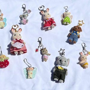 May include: A collection of miniature animal keychains, including bunnies, a koala, a bear, a dog, and an elephant. The keychains feature various outfits and accessories, such as dresses, overalls, and wings. Each keychain has a silver clasp.
