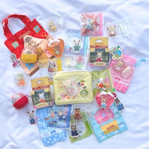May include: A collection of Calico Critters toys and accessories, including miniature animal figures, packaged in boxes, bags, and clear plastic containers. The items are arranged on a white surface, with a red tote bag and a yellow cosmetic bag.