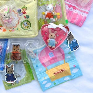May include: A collection of Sylvanian Families toys, including animal figurines and accessories. The image features a variety of packaged sets with different characters, a heart-shaped container, and a bag with a hot air balloon design. The color palette includes pastels and bright colors.