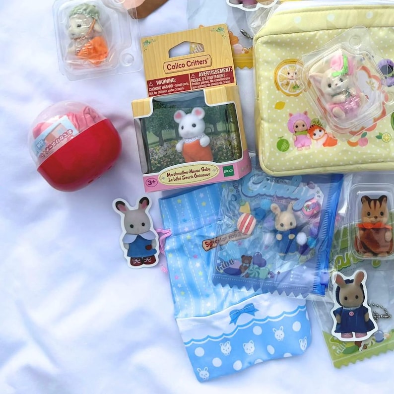 May include: A collection of Calico Critters toys and accessories, including miniature animal figures, a red and white capsule toy, and a light yellow pouch with a lemon and character design. The toys are packaged in clear plastic and cardboard boxes.