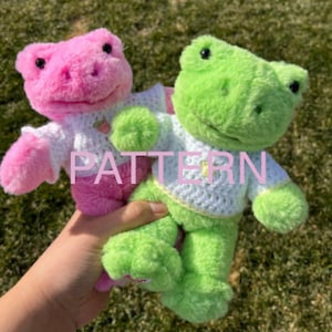 May include: Two plush frog toys wearing crocheted sweaters. The frog on the left is pink and the frog on the right is green. Both frogs have white sweaters with yellow accents.
